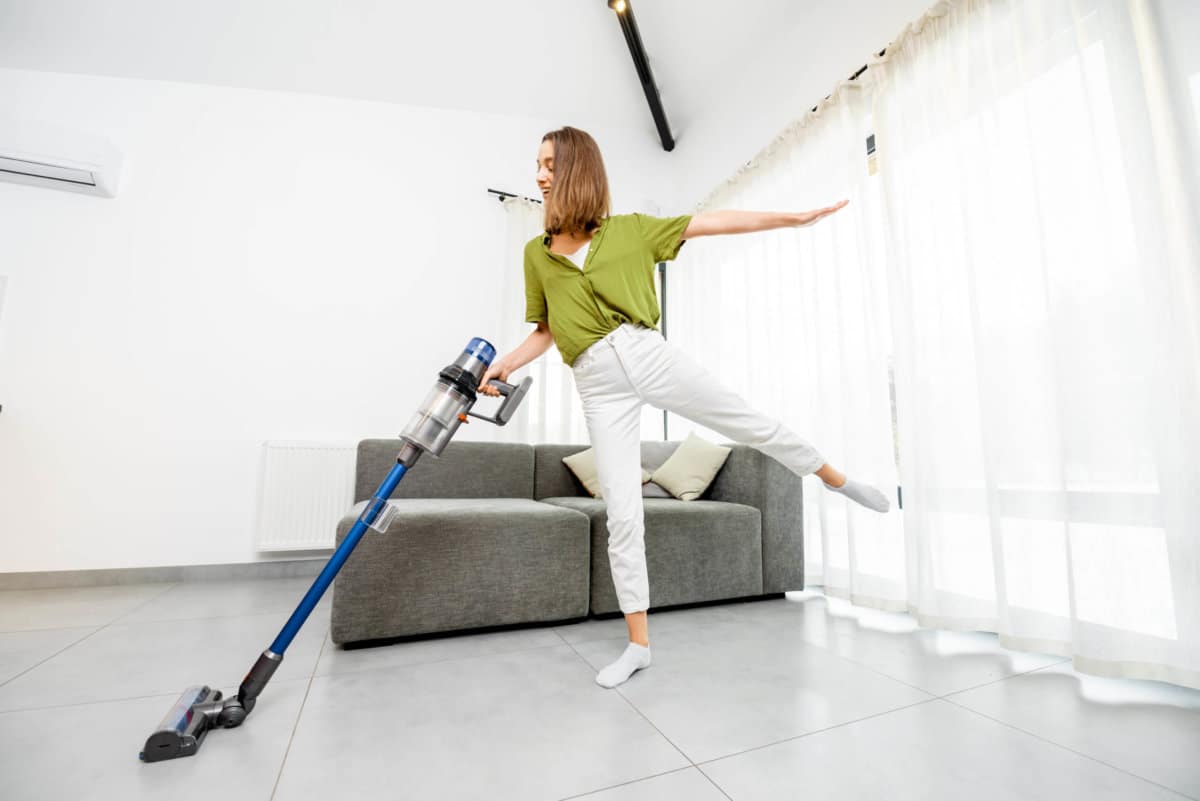 Young woman enjoying housework, cleaning floor with cordless vacuum cleaner in the modern white living room. Concept of easy cleaning with a wireless vacuum cleaner